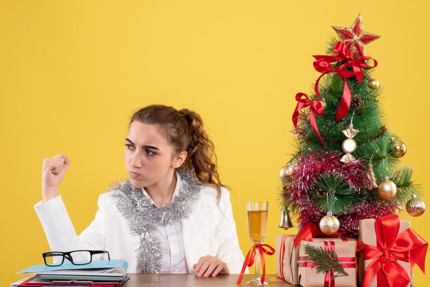 Vue de face femme médecin assis derrière la table avec des cadeaux de Noël et arbre sur un bureau jaune