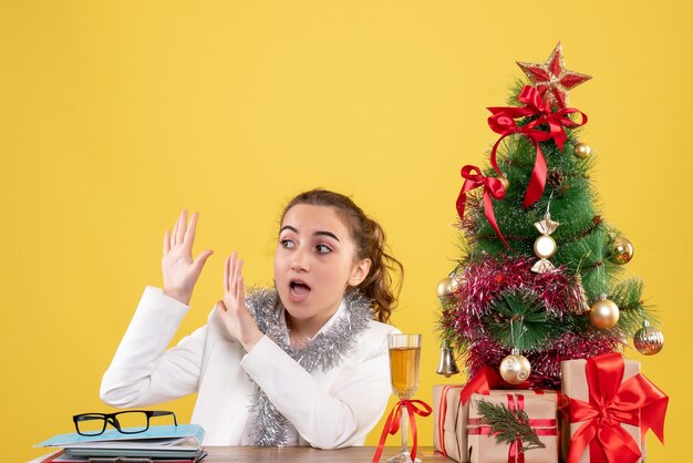 Vue de face femme médecin assis derrière la table avec des cadeaux de Noël et arbre sur fond jaune