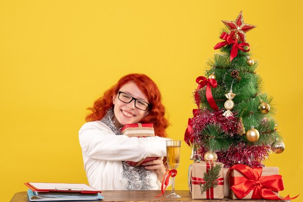 Vue de face femme médecin assis derrière la table avec des cadeaux de Noël sur un bureau jaune avec arbre de Noël et coffrets cadeaux