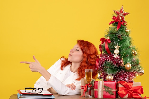 Vue de face femme médecin assis derrière la table avec des cadeaux de Noël sur un bureau jaune