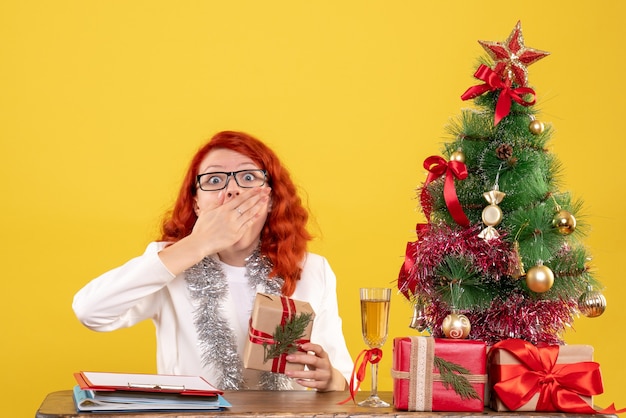 Vue de face femme médecin assis derrière la table avec des cadeaux de Noël choqué sur fond jaune