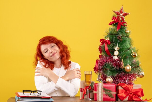 Vue de face femme médecin assis derrière la table avec des cadeaux de Noël sur fond jaune avec arbre de Noël et coffrets cadeaux