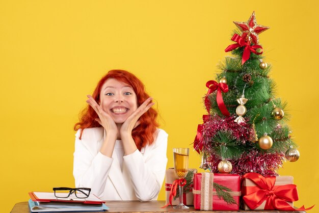 Vue de face femme médecin assis derrière la table avec des cadeaux de Noël sur fond jaune