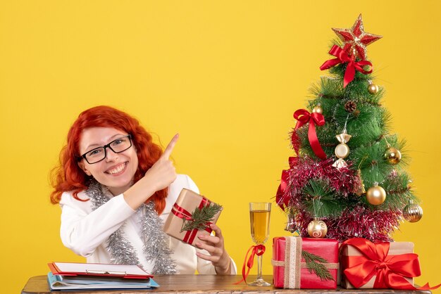 Vue de face femme médecin assis derrière la table avec des cadeaux de Noël sur fond jaune