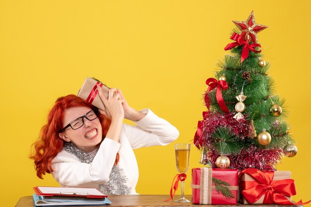 Vue de face femme médecin assis derrière la table avec des cadeaux de Noël sur fond jaune