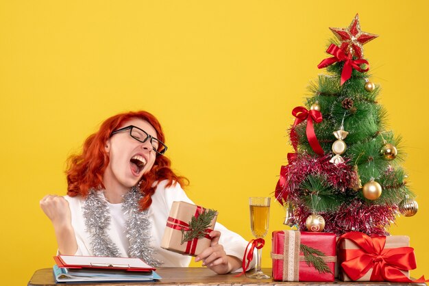 Vue de face femme médecin assis derrière la table avec des cadeaux de Noël sur fond jaune