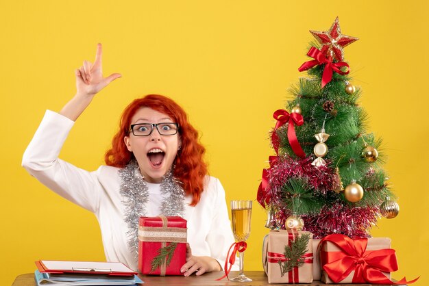 Vue de face femme médecin assis derrière la table avec des cadeaux de Noël sur fond jaune