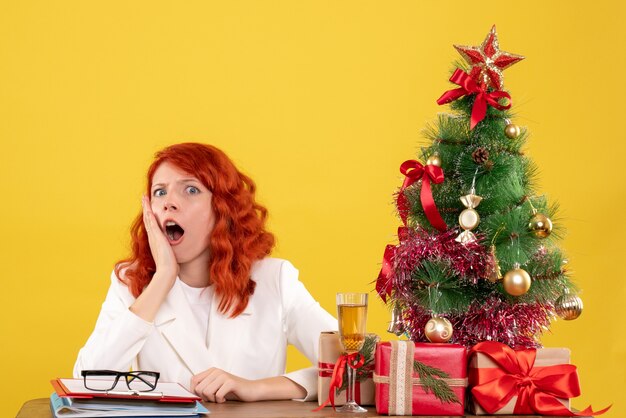 Vue de face femme médecin assis derrière la table avec des cadeaux de Noël sur fond jaune