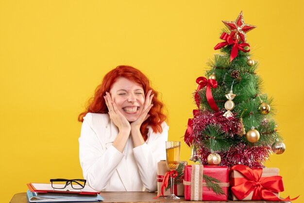 Vue de face femme médecin assis derrière la table avec des cadeaux de Noël sur fond jaune