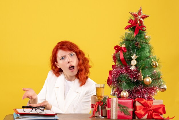 Vue de face femme médecin assis derrière la table avec des cadeaux de Noël sur fond jaune