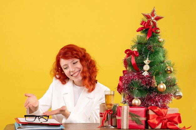 Vue de face femme médecin assis derrière la table avec des cadeaux de Noël sur fond jaune