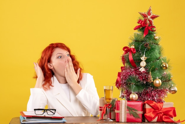 Photo gratuite vue de face femme médecin assis derrière la table avec des cadeaux de noël surpris sur fond jaune