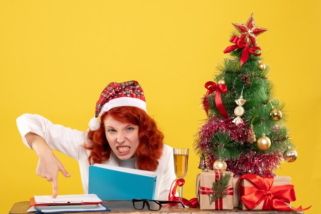 Vue de face femme médecin assis derrière la table avec des documents dans ses mains sur fond jaune avec arbre de Noël et coffrets cadeaux