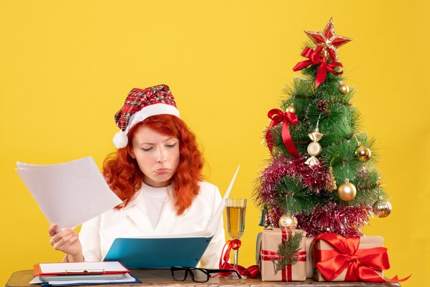 Vue de face femme médecin assis derrière la table avec des documents sur fond jaune avec arbre de Noël et coffrets cadeaux