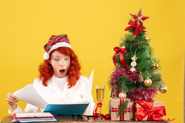 Vue de face femme médecin assis derrière la table de lecture de documents sur fond jaune avec arbre de Noël et coffrets cadeaux