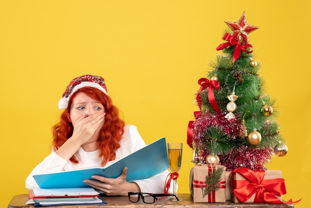 Vue de face femme médecin assis derrière la table de lecture de documents sur fond jaune avec arbre de Noël et coffrets cadeaux