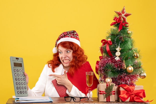 Vue de face femme médecin assis derrière la table et tenant la calculatrice sur fond jaune avec arbre de Noël et coffrets cadeaux