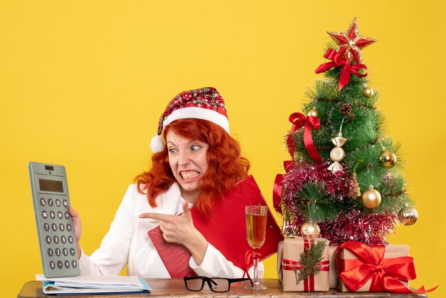 Vue de face femme médecin assis derrière la table et tenant la calculatrice sur fond jaune avec arbre de Noël et coffrets cadeaux