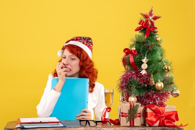 Vue de face femme médecin assis derrière la table et tenant des documents sur fond jaune avec arbre de Noël et coffrets cadeaux