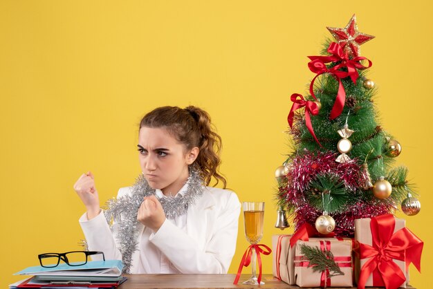 Vue de face femme médecin assis derrière la table avec visage en colère sur fond jaune avec arbre de Noël et coffrets cadeaux