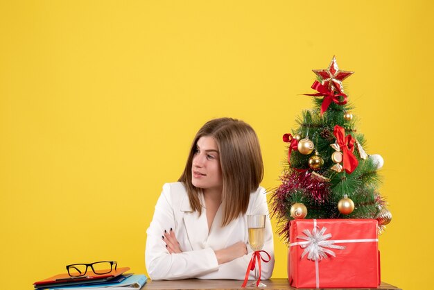 Vue de face femme médecin assis devant sa table sur un bureau jaune avec arbre de Noël et coffrets cadeaux