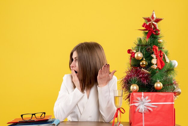 Vue de face femme médecin assis devant sa table sur fond jaune avec arbre de Noël et coffrets cadeaux