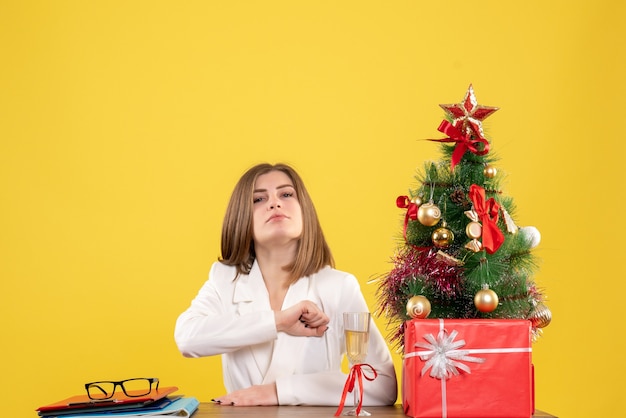 Vue de face femme médecin assis devant sa table sur fond jaune avec arbre de Noël et coffrets cadeaux