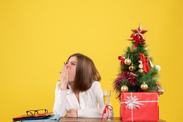 Vue de face femme médecin assis devant sa table sur fond jaune avec arbre de Noël et coffrets cadeaux