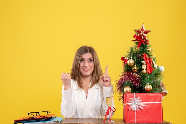 Vue de face femme médecin assis devant sa table sur fond jaune avec arbre de Noël et coffrets cadeaux