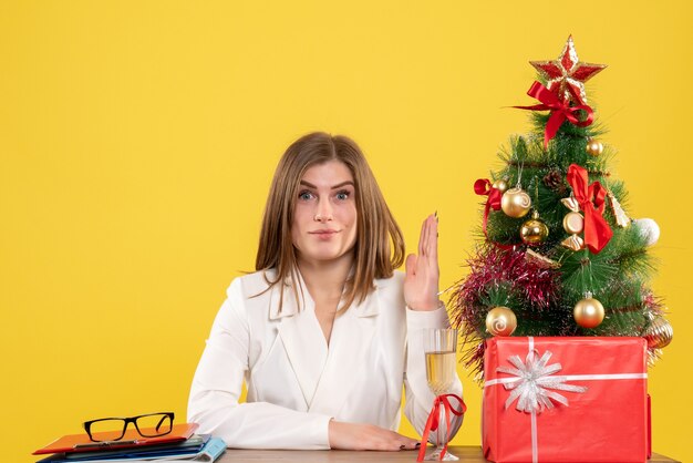 Vue de face femme médecin assis devant sa table sur fond jaune avec arbre de Noël et coffrets cadeaux