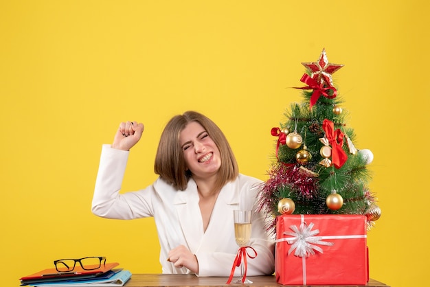 Vue de face femme médecin assis devant sa table sur fond jaune avec arbre de Noël et coffrets cadeaux