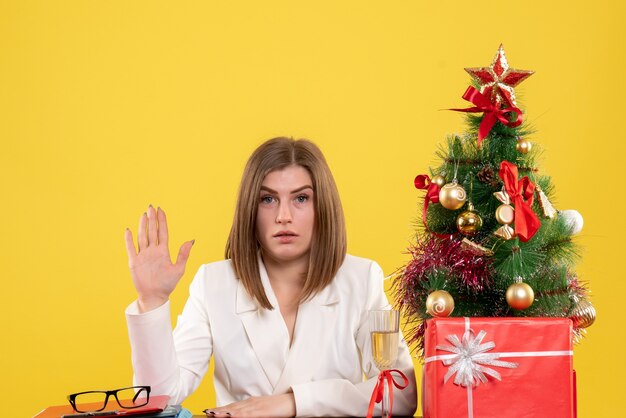 Vue de face femme médecin assis devant sa table sur fond jaune avec arbre de Noël et coffrets cadeaux