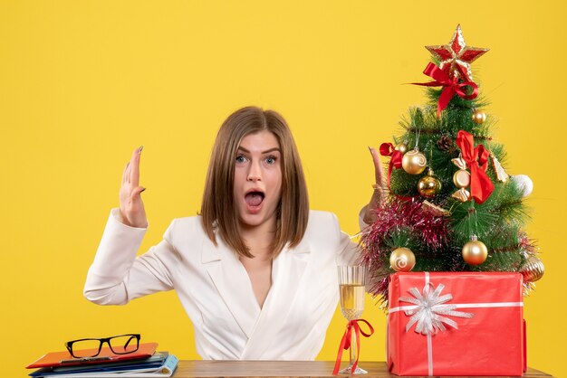 Vue de face femme médecin assis devant sa table sur fond jaune avec arbre de Noël et coffrets cadeaux