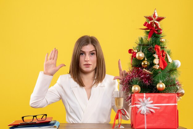 Vue de face femme médecin assis devant sa table sur fond jaune avec arbre de Noël et coffrets cadeaux