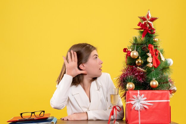 Vue de face femme médecin assis devant sa table sur fond jaune avec arbre de Noël et coffrets cadeaux