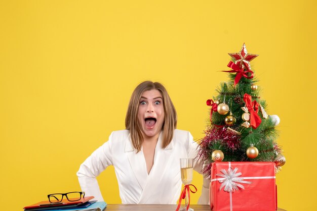 Vue de face femme médecin assis devant sa table sur fond jaune avec arbre de Noël et coffrets cadeaux