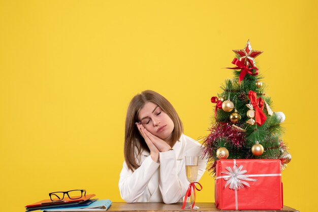 Vue de face femme médecin assis devant sa table sur fond jaune avec arbre de Noël et coffrets cadeaux