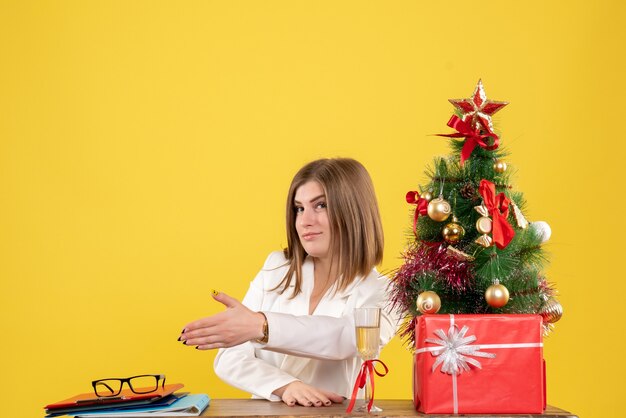 Vue de face femme médecin assis devant sa table se serrant la main sur fond jaune avec arbre de Noël et coffrets cadeaux