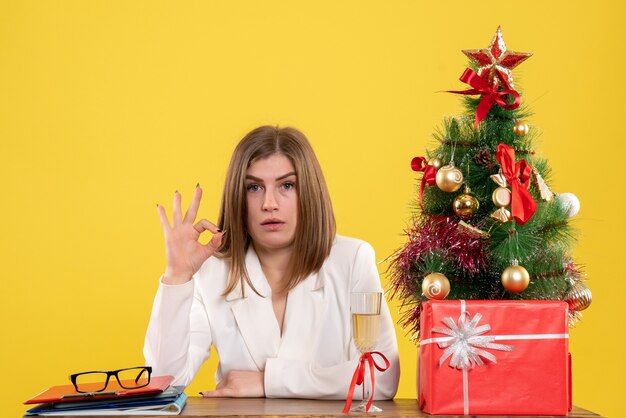 Vue de face femme médecin assis devant sa table sur un sol jaune avec arbre de Noël et coffrets cadeaux