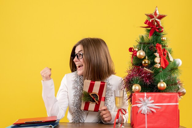 Vue de face femme médecin assis devant sa table tenant présent sur fond jaune avec arbre de Noël et coffrets cadeaux