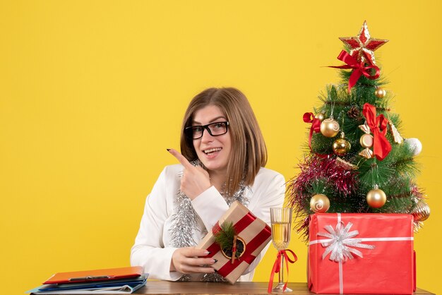 Vue de face femme médecin assis devant sa table tenant présent sur fond jaune avec arbre de Noël et coffrets cadeaux