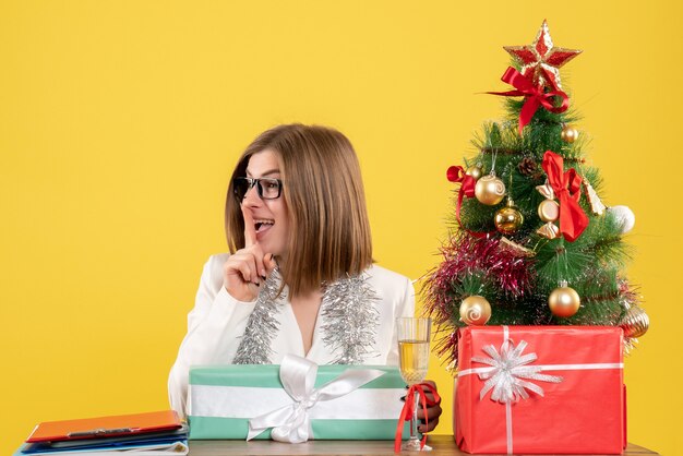 Vue de face femme médecin assis devant la table avec des cadeaux et arbre sur fond jaune avec arbre de Noël et coffrets cadeaux