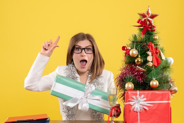 Vue de face femme médecin assis devant la table avec des cadeaux et arbre sur fond jaune avec arbre de Noël et coffrets cadeaux