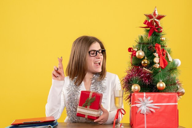 Vue de face femme médecin assis devant la table avec des cadeaux et arbre sur fond jaune avec arbre de Noël et coffrets cadeaux