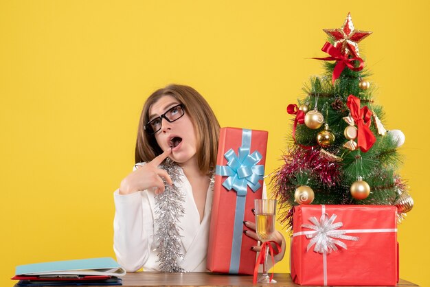 Vue de face femme médecin assis devant la table avec des cadeaux et arbre sur fond jaune avec arbre de Noël et coffrets cadeaux