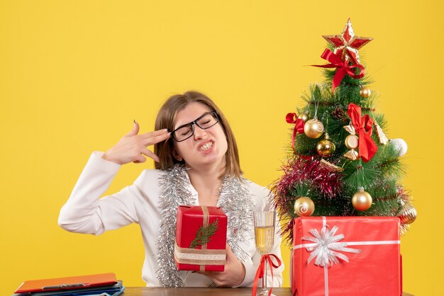 Vue de face femme médecin assis devant la table avec des cadeaux et arbre sur fond jaune avec arbre de Noël et coffrets cadeaux