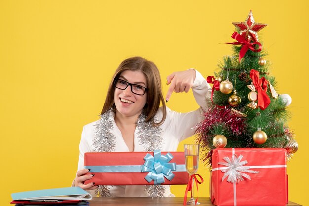 Vue de face femme médecin assis devant la table avec des cadeaux et arbre sur fond jaune avec arbre de Noël et coffrets cadeaux