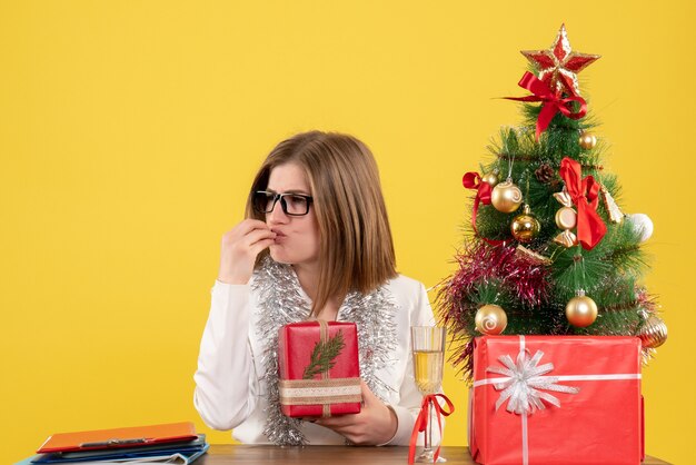 Vue de face femme médecin assis en face de la table avec des cadeaux et arbre sur un bureau jaune