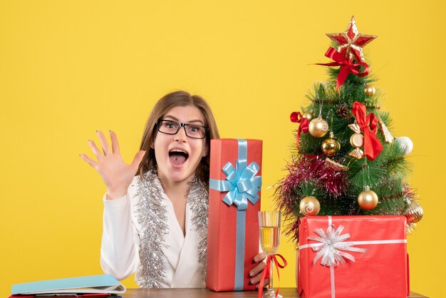 Vue de face femme médecin assis en face de la table avec des cadeaux et arbre sur fond jaune avec arbre de Noël et coffrets cadeaux