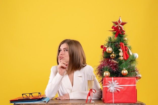 Vue de face femme médecin assis en face de table avec des cadeaux de Noël et arbre sur fond jaune